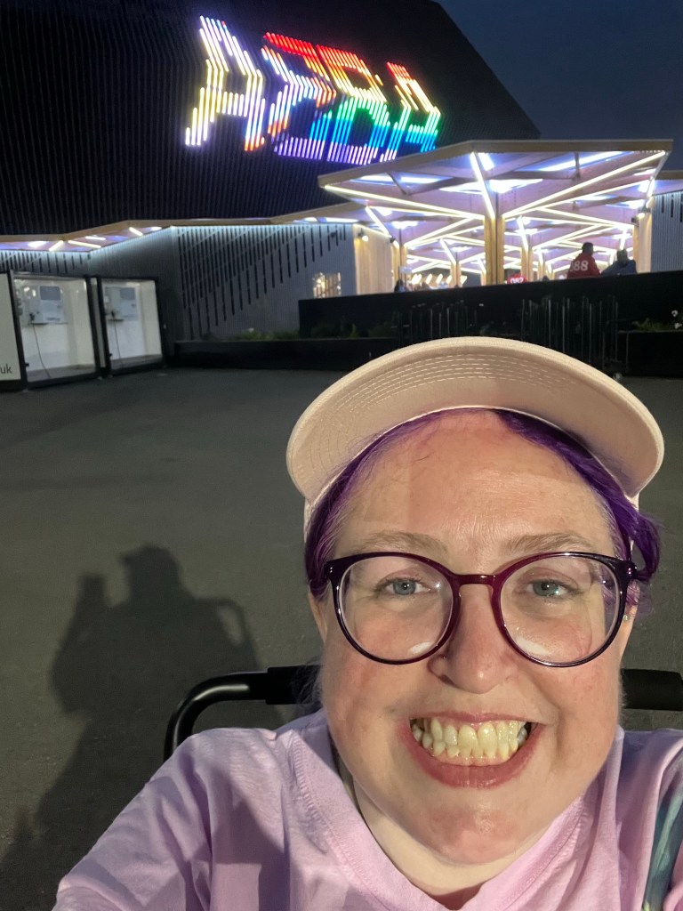Vikki has taken a selfie in front of the arena. ABBA is lit up in progress pride flag colours. (Rainbow, trans and intersex). She smiles at the camera and is wearing a cap. 
