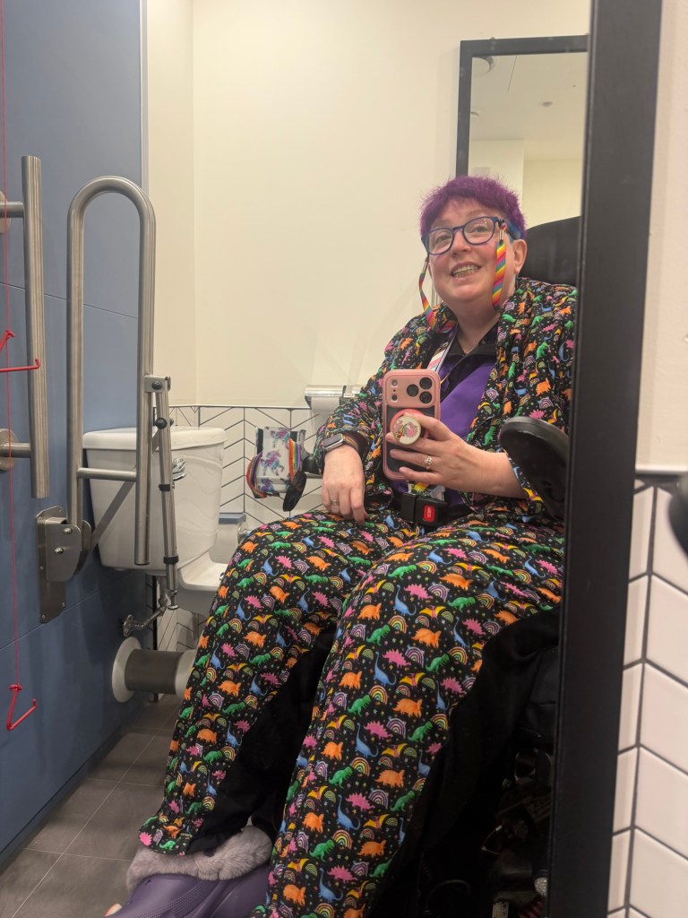 Vikki sits in their powerchair taking a photo in a mirror in an accessible loo. They have short hair that is purple and blue and wear blue glasses with a rainbow cord to not lose them. They are wearing a multicoloured suit covered in dinosaurs. 